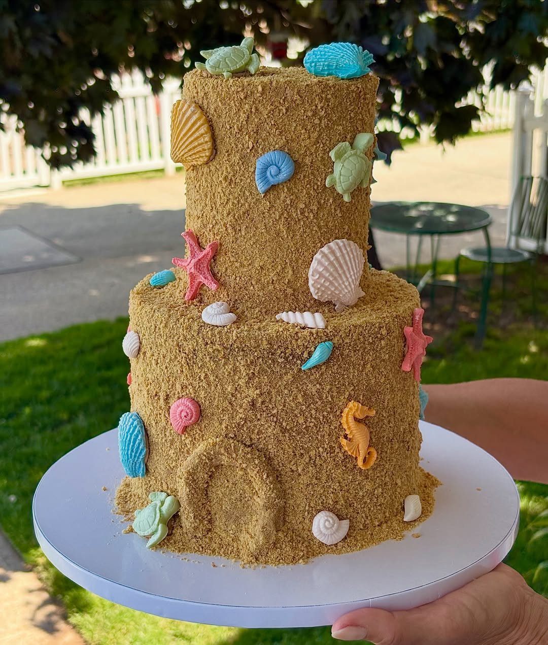 Two-tiered sand-textured cake decorated with colorful edible seashells, starfish, and sea turtles.